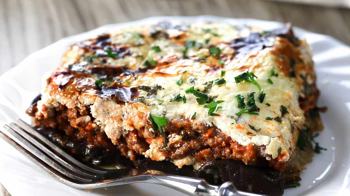 A slice of Moussaka on a white plate and a fork next to it.