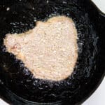 A breaded pork chop beginning to fry in oil in a cast-iron skillet.