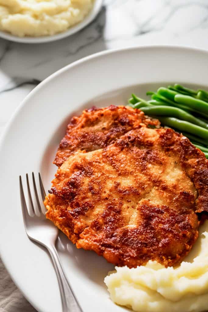 A crispy panko-breaded pork chop served with mashed potatoes and green beans on a white plate.