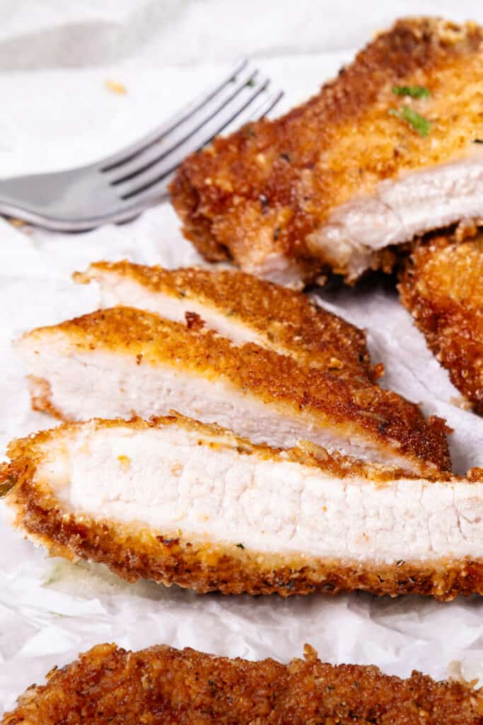 Sliced panko-breaded pork chop showing a juicy interior with a crispy crust, beside a fork on a white paper.