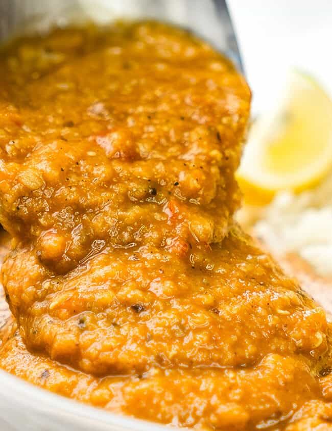 A close up image of a spoon serving up vibrant red lentil dal from a bowl, with fluffy white rice and lemon wedges in the background.