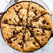 A wide-angle shot of a freshly baked chocolate chunk Pizookie skillet cookie, cut into wedges, with a wooden spatula, all resting on a wire cooling rack over a marble background.