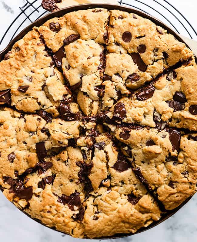 A wide-angle shot of a freshly baked chocolate chunk Pizookie skillet cookie, cut into wedges, with a wooden spatula, all resting on a wire cooling rack over a marble background.