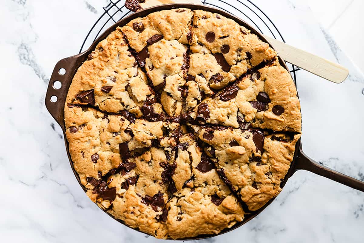 A wide-angle shot of a freshly baked chocolate chunk Pizookie skillet cookie, cut into wedges, with a wooden spatula, all resting on a wire cooling rack over a marble background.