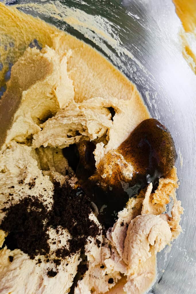 Overhead view of cookie dough in a mixing bowl with espresso powder and vanilla extract being added, ready for mixing.