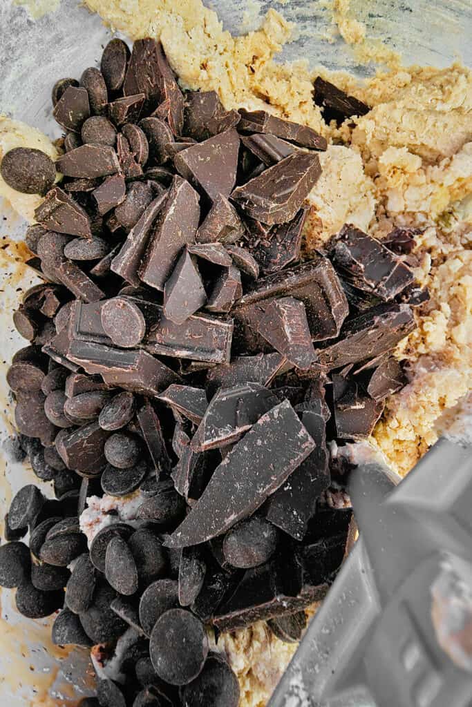 A pile of dark chocolate chunks and chocolate chips on top of cookie dough in a mixing bowl, ready to be stirred in.