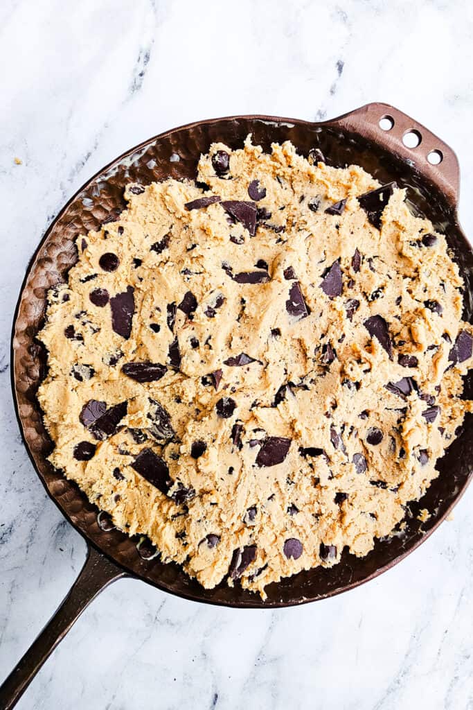 Overhead image of chocolate chip cookie dough spread evenly in a round cast-iron skillet, placed on a white marble surface.
