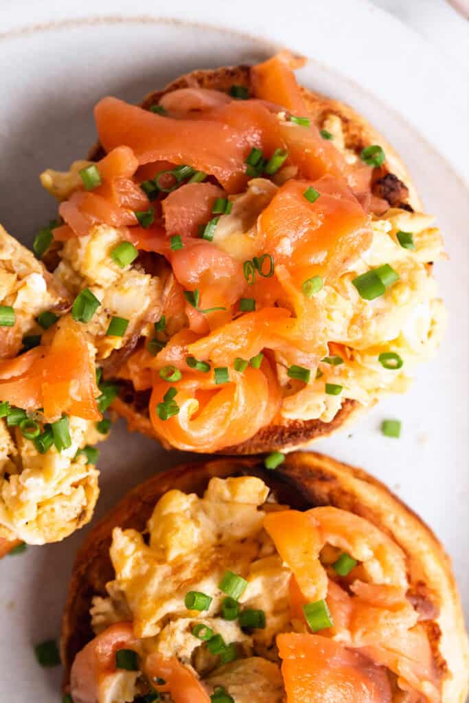 A plate of breakfast food with two halves of an English muffin topped with scrambled eggs and smoked salmon.