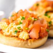 Two toasted English muffins topped with scrambled eggs and smoked salmon on a white plate.