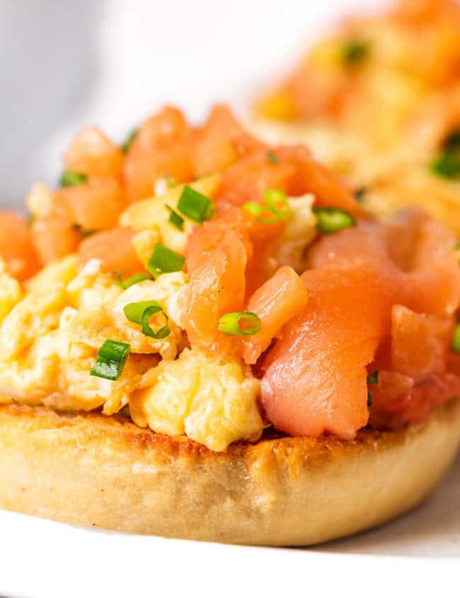 Two toasted English muffins topped with scrambled eggs and smoked salmon on a white plate.