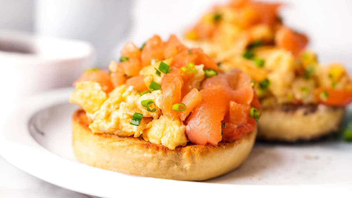 Two toasted English muffins topped with scrambled eggs and smoked salmon on a white plate.