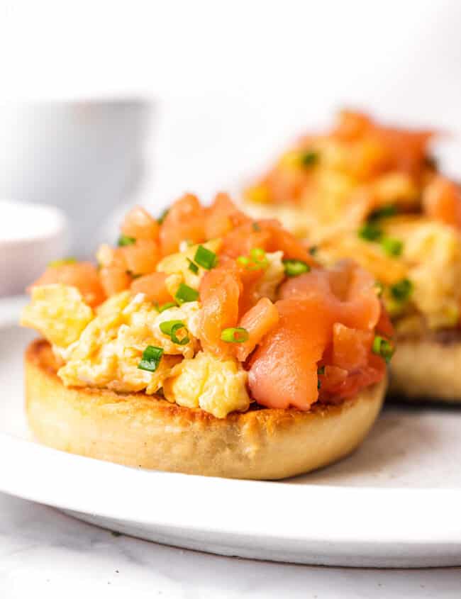 Two toasted English muffins topped with scrambled eggs and smoked salmon on a white plate.