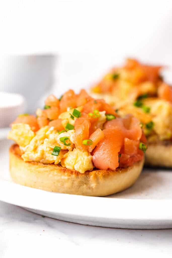 Two toasted English muffins topped with scrambled eggs and smoked salmon on a white plate.