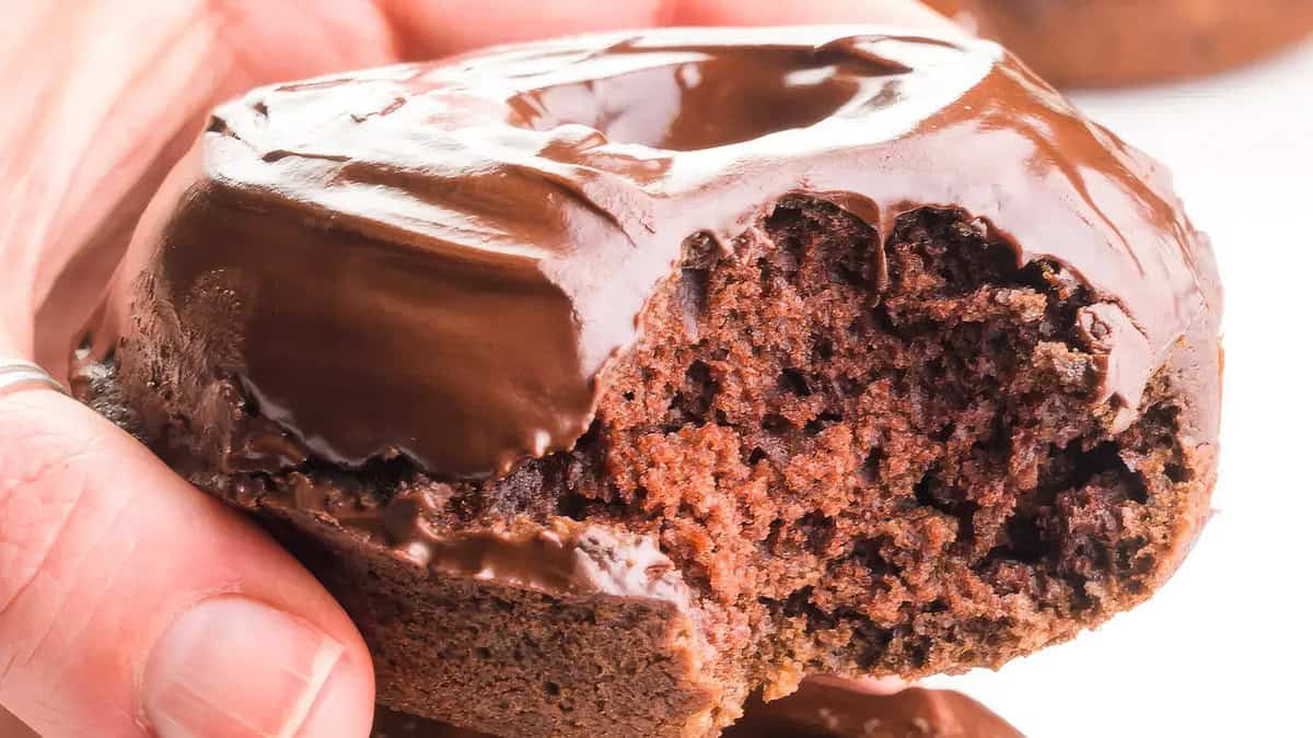 A close-up photo of a chocolate donut with a single bite taken out, revealing a fluffy interior. The donut is in someone's hand.