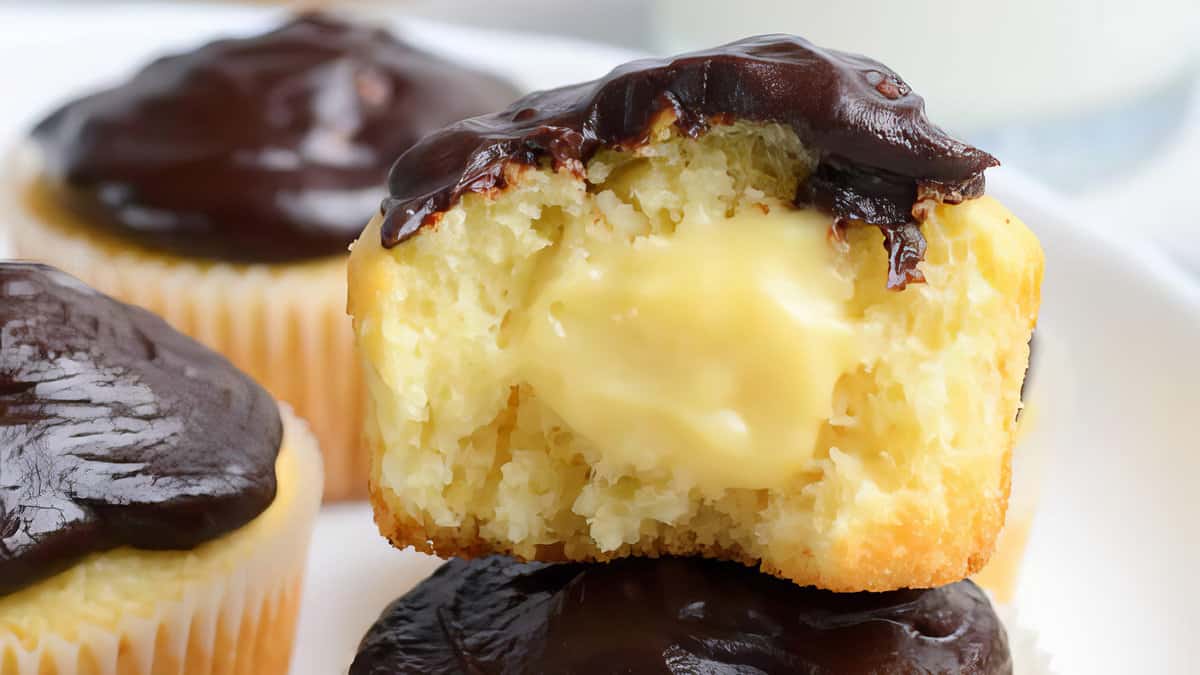 A Boston cream pie cupcake with a bite taken out, revealing a creamy vanilla filling. The cupcake is topped with chocolate ganache and sits on a white plate.