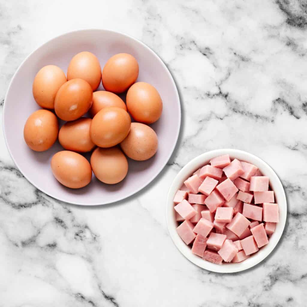 a photo of a bowl of 12 eggs and a bowl of diced ham on a marble surface.