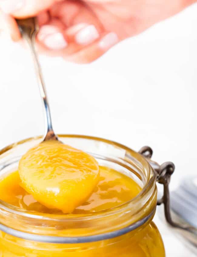 A hand holding a spoonful of lemon curd over a jar of lemon curd.