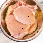 Pork chops marinating in a clear brine with peppercorns in a glass bowl, atop a striped kitchen towel.