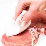 A hand with a pink manicure patting a raw pork chop dry with a paper towel on a white surface.