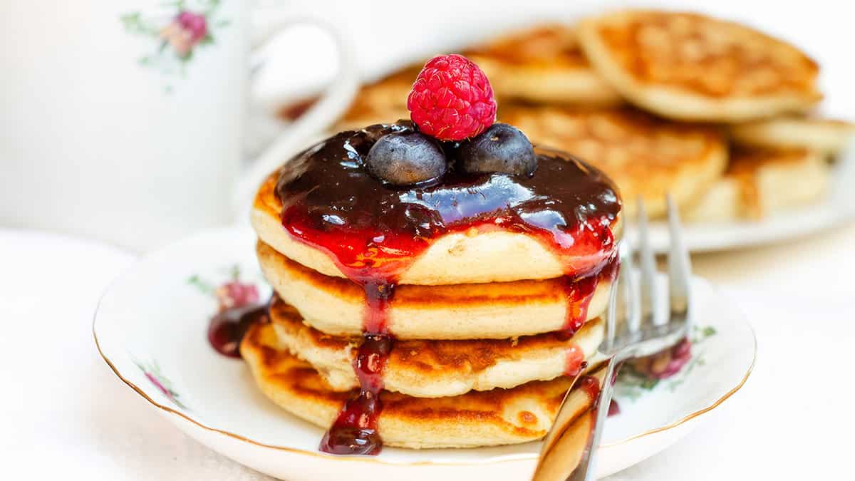 A stack of Scotch pancakes topped with a glossy berry sauce and fresh berries, with a cup of tea and extra pancakes in the background, all on a white tablecloth.