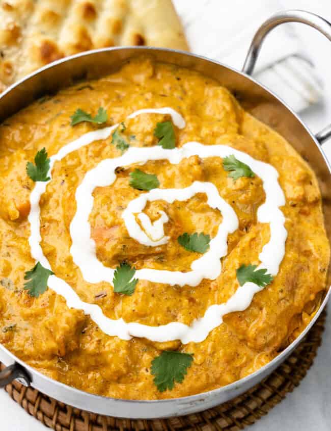 butter chicken curry served in a metal bowl, beautifully garnished with swirls of white yogurt and fresh green cilantro leaves on top.