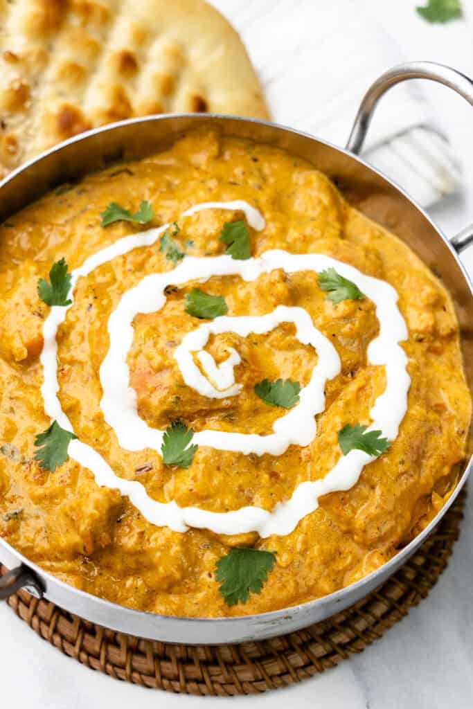  butter chicken curry served in a metal bowl, beautifully garnished with swirls of white yogurt and fresh green cilantro leaves on top.