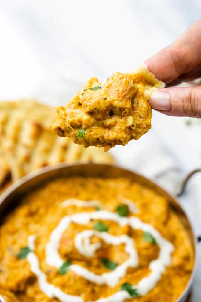  A close-up image capturing a piece of naan bread being dipped into creamy butter chicken curry. The curry, visible in the background.