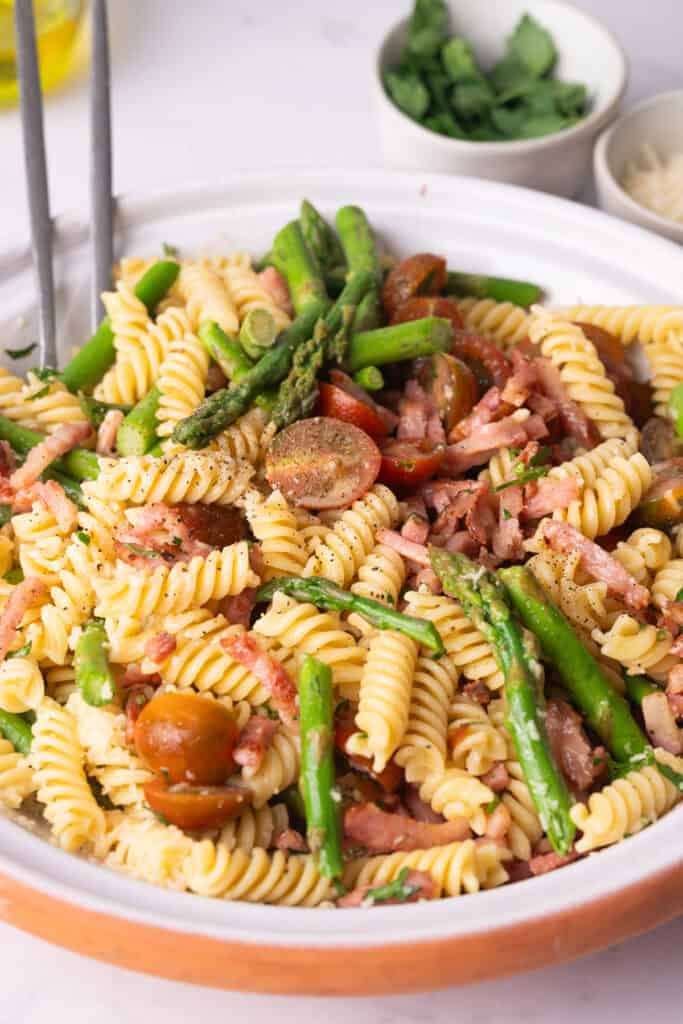 Bowl of bacon and asparagus pasta salad with fusilli pasta, cherry tomatoes, and grated Parmesan, served on a white plate with a small bowl of fresh parsley in the background.