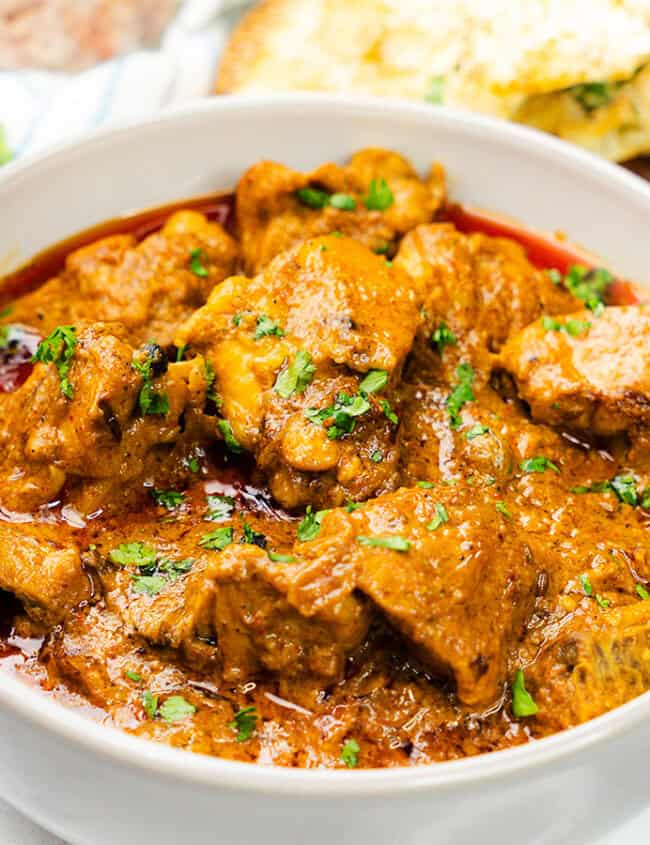 A bowl filled with Chicken Korma, garnished with fresh cilantro, sits beside garlic naan on a woven mat.