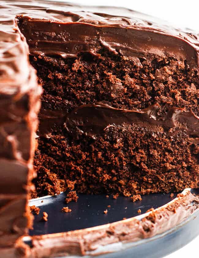 A devil's food cake with a slice removed, displaying the rich layers of cake and smooth chocolate frosting, on a white plate atop a dark slate board, against a marble background.