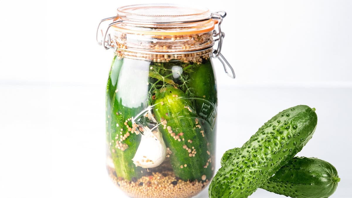 A jar of pickled cucumbers with garlic, mustard seeds, and dill fronds on a wooden table next to two whole cucumbers.