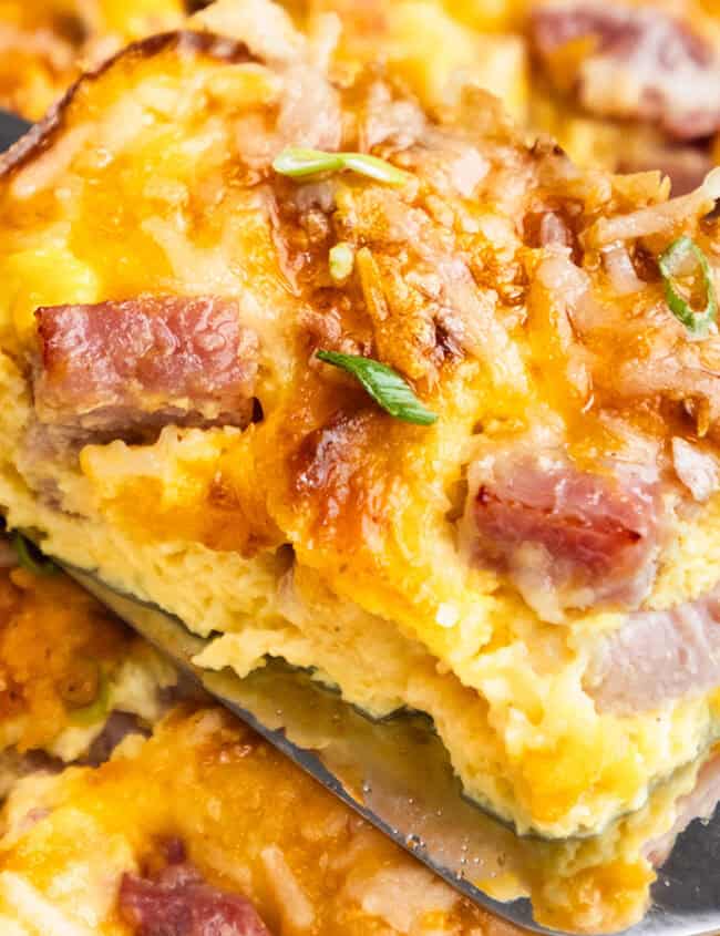 A slice of ham and cheese casserole being lifted with a spatula, showing the layered interior, garnished with herbs.