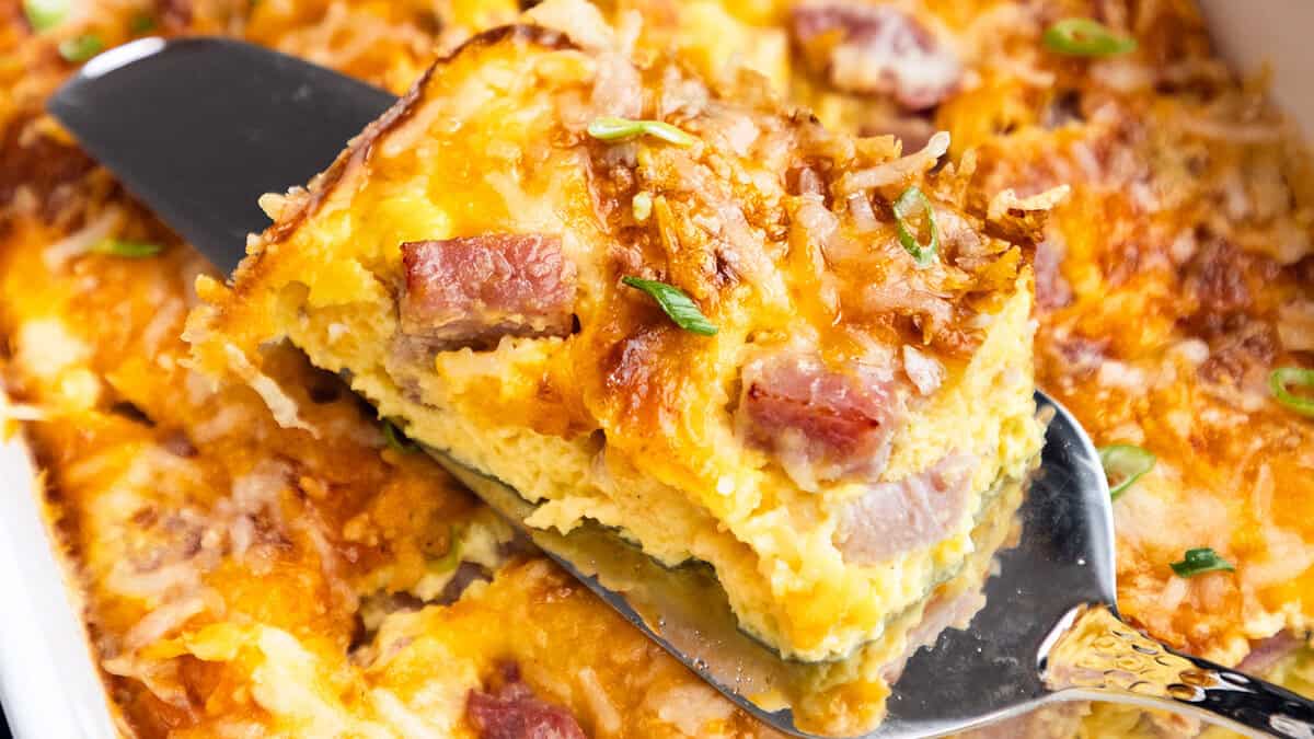 A slice of ham and cheese casserole being lifted with a spatula, showing the layered interior, garnished with herbs.