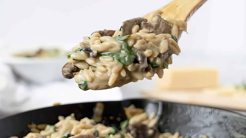 A wooden spoon filled with orzo pasta and chopped mushrooms in a stainless steel skillet.
