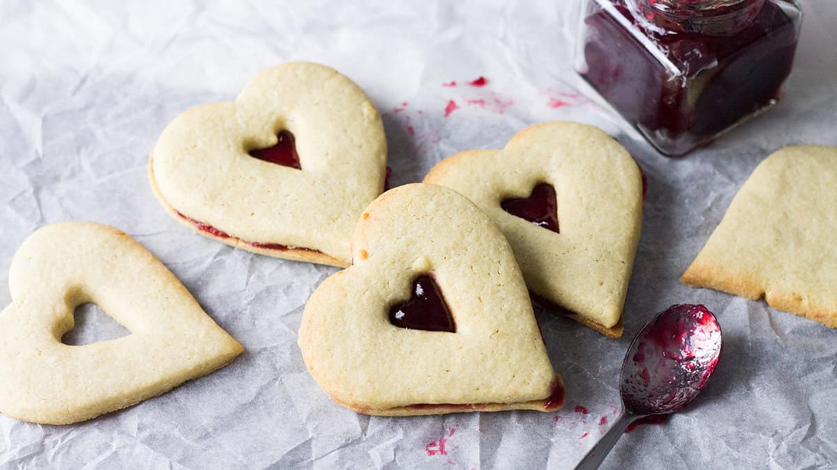 Heart-shaped butter cookies with raspberry jam filling on parchment paper with a spoon smeared with jam
