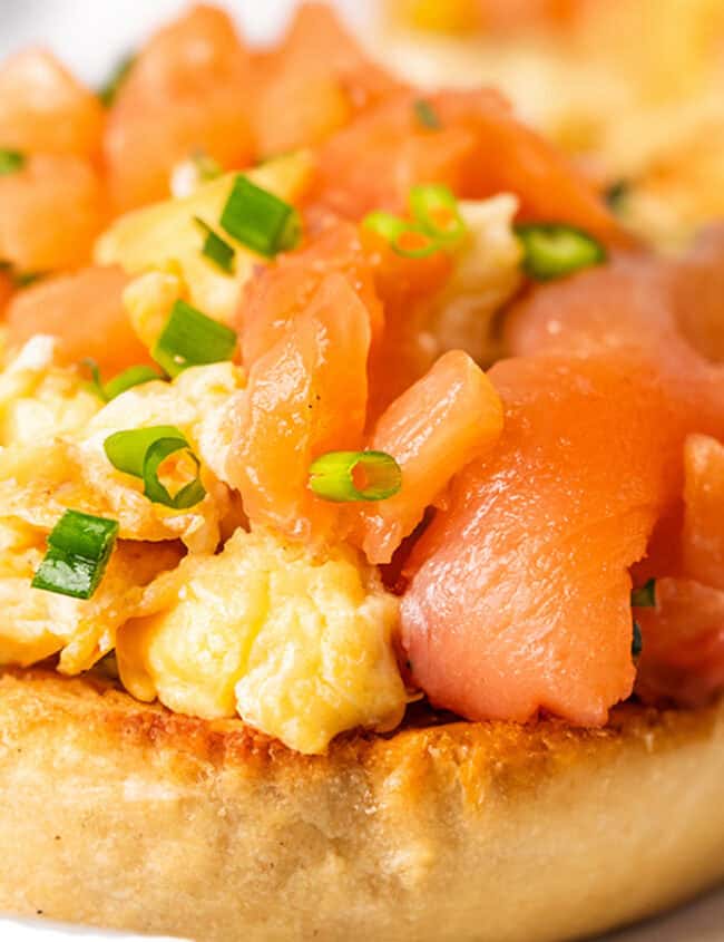 Two toasted English muffins topped with scrambled eggs and smoked salmon on a white plate.