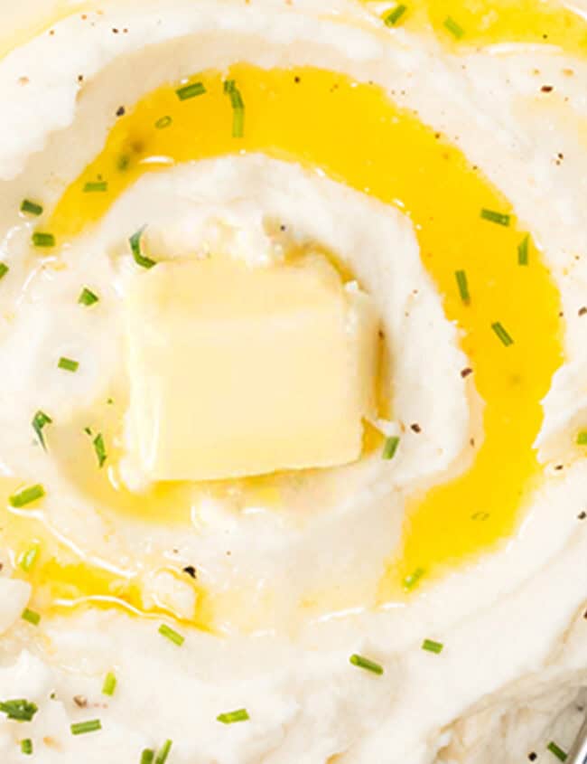 A close up photo taken from above of a bowl of Sour Cream Mashed Potatoes with melted butter and chives with a serving spoon.