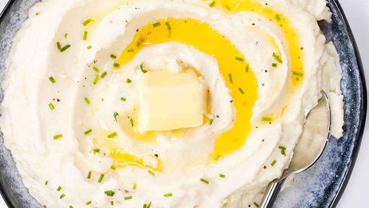 A close up photo taken from above of a bowl of Sour Cream Mashed Potatoes with melted butter and chives with a serving spoon.