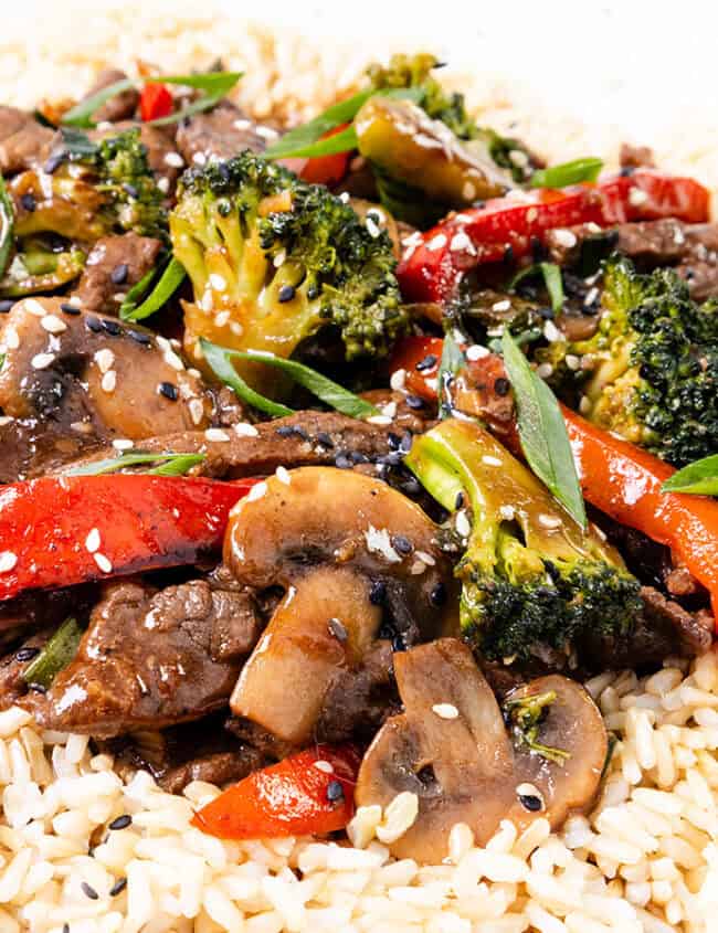 A steak stir fry with broccoli, red bell peppers, and mushrooms served over white rice, garnished with sesame seeds and accompanied by black chopsticks.