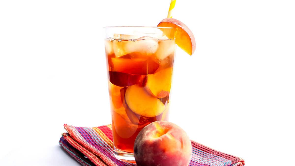 A glass of Sweet Peach Iced Tea with ice and a straw and a fresh wedge of peach on the glass.