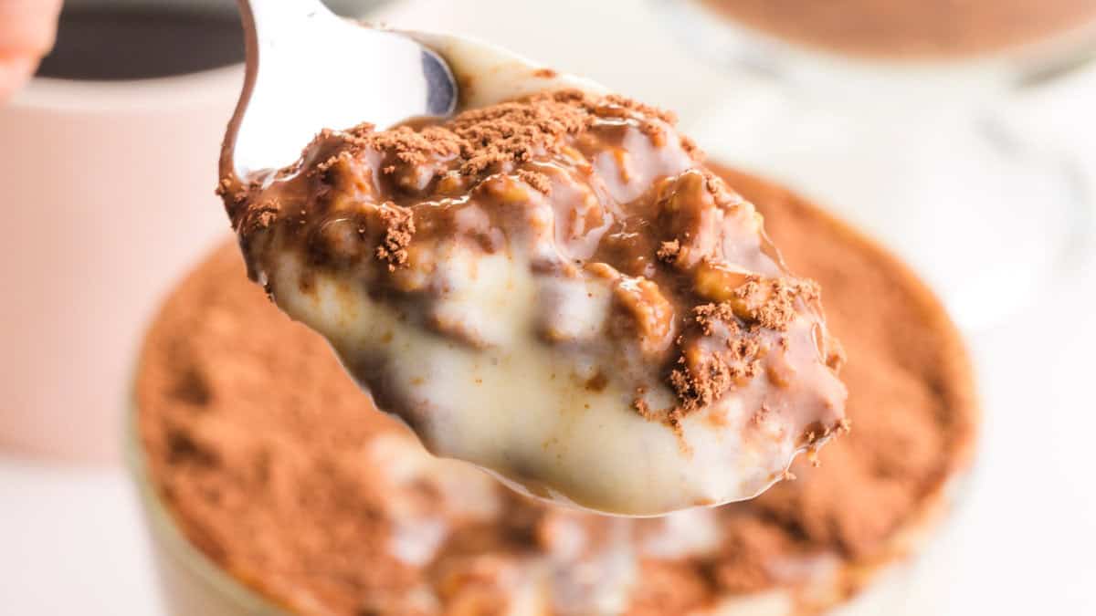 A close-up view of a glass jar filled with layered Tiramisu Overnight Oats. The oats appear light brown and are layered with dark cocoa powder and coffee grounds.