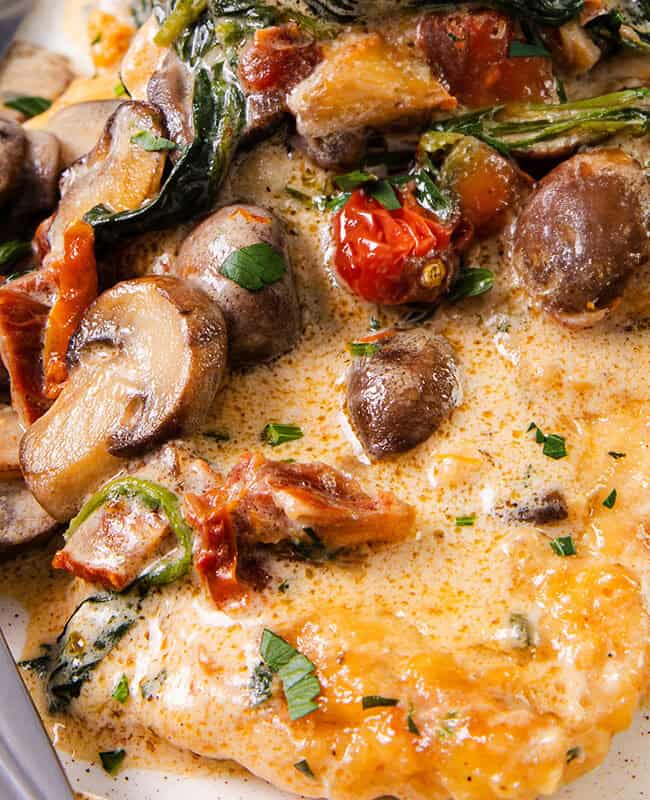 A landscape image of a plate of creamy Tuscan chicken with sun-dried tomatoes, spinach, and mushrooms, served with a fork and knife.