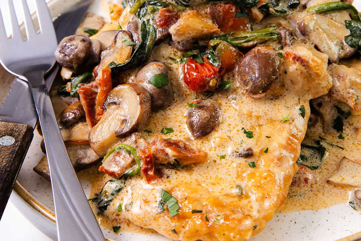 A landscape image of a plate of creamy Tuscan chicken with sun-dried tomatoes, spinach, and mushrooms, served with a fork and knife.