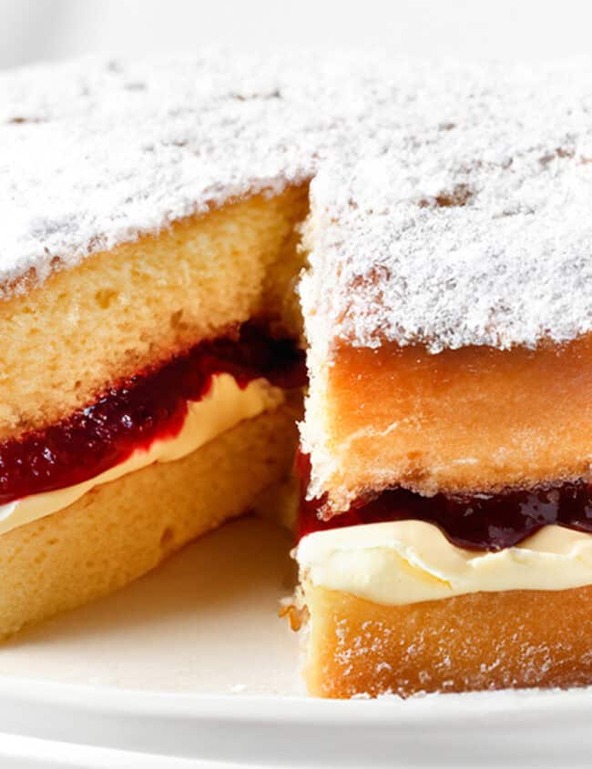 A close up image of Victoria sponge cake on a cake stand with a slice cut out of it.