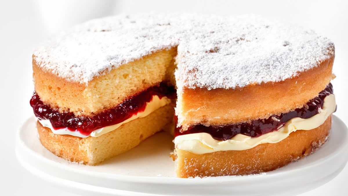 A close up image of Victoria sponge cake on a cake stand with a slice cut out of it.