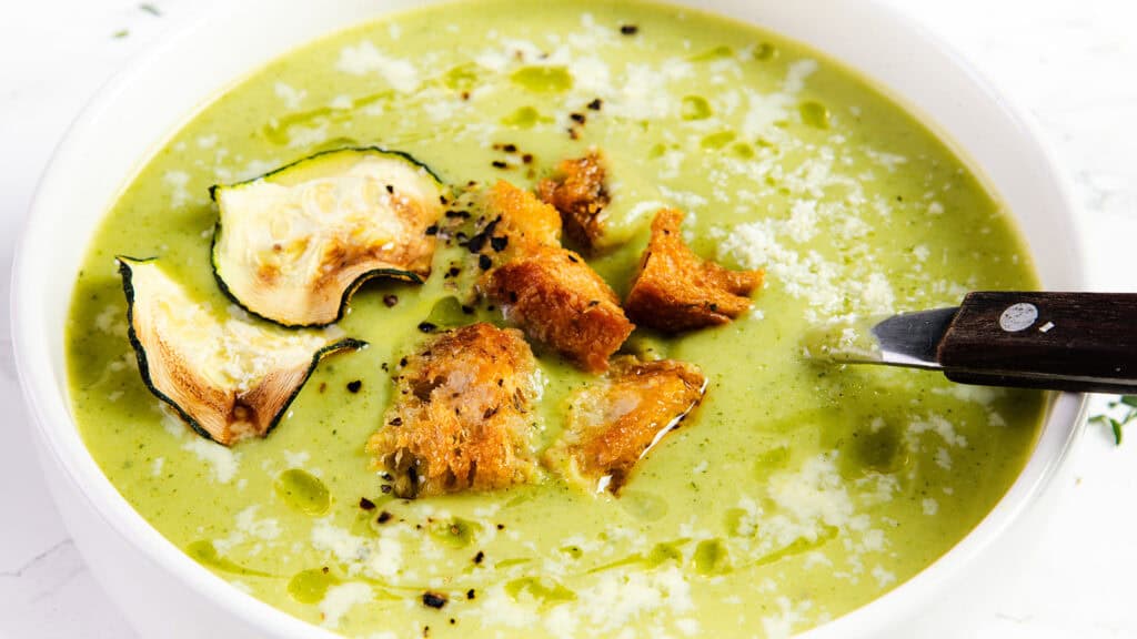 A close-up photo of a bowl of creamy zucchini soup. The soup is a light green color and garnished with a swirl of heavy cream and a sprinkle of pepper.