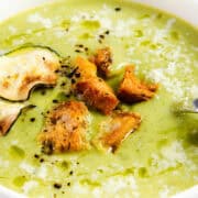 A close-up photo of a bowl of creamy zucchini soup. The soup is a light green color and garnished with a swirl of heavy cream and a sprinkle of pepper.