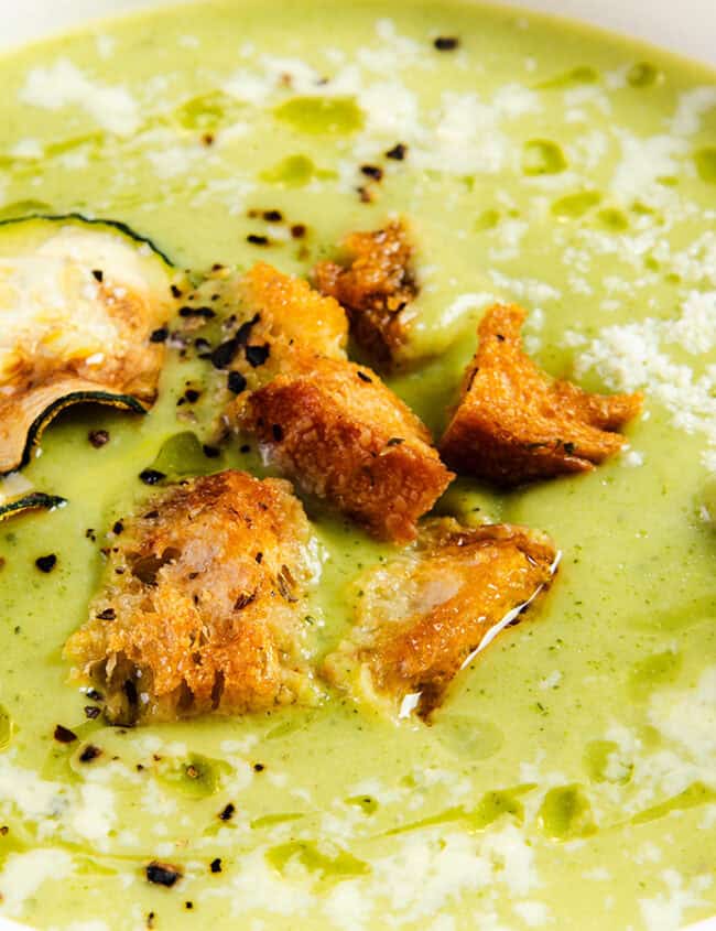 A close-up photo of a bowl of creamy zucchini soup. The soup is a light green color and garnished with a swirl of heavy cream and a sprinkle of pepper.