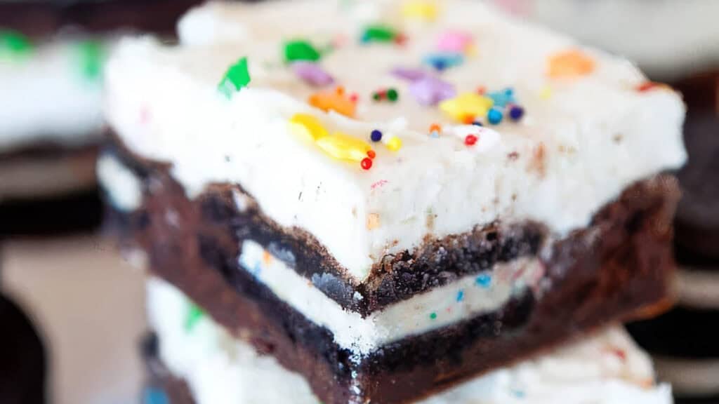 A close-up of a slice of oreo cream cheese brownie. The brownie has a thick layer of cream cheese frosting in the center and is topped with crushed Oreos and white chocolate chips.
