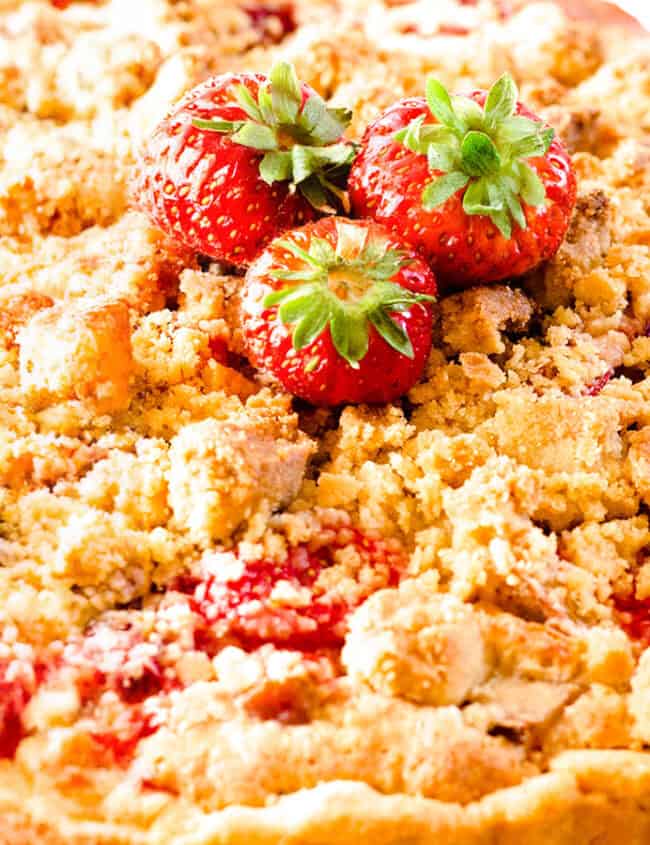 A close up of a Strawberry Rhubarb Pie with a crumb topping on a serving plate topped with strawberries.